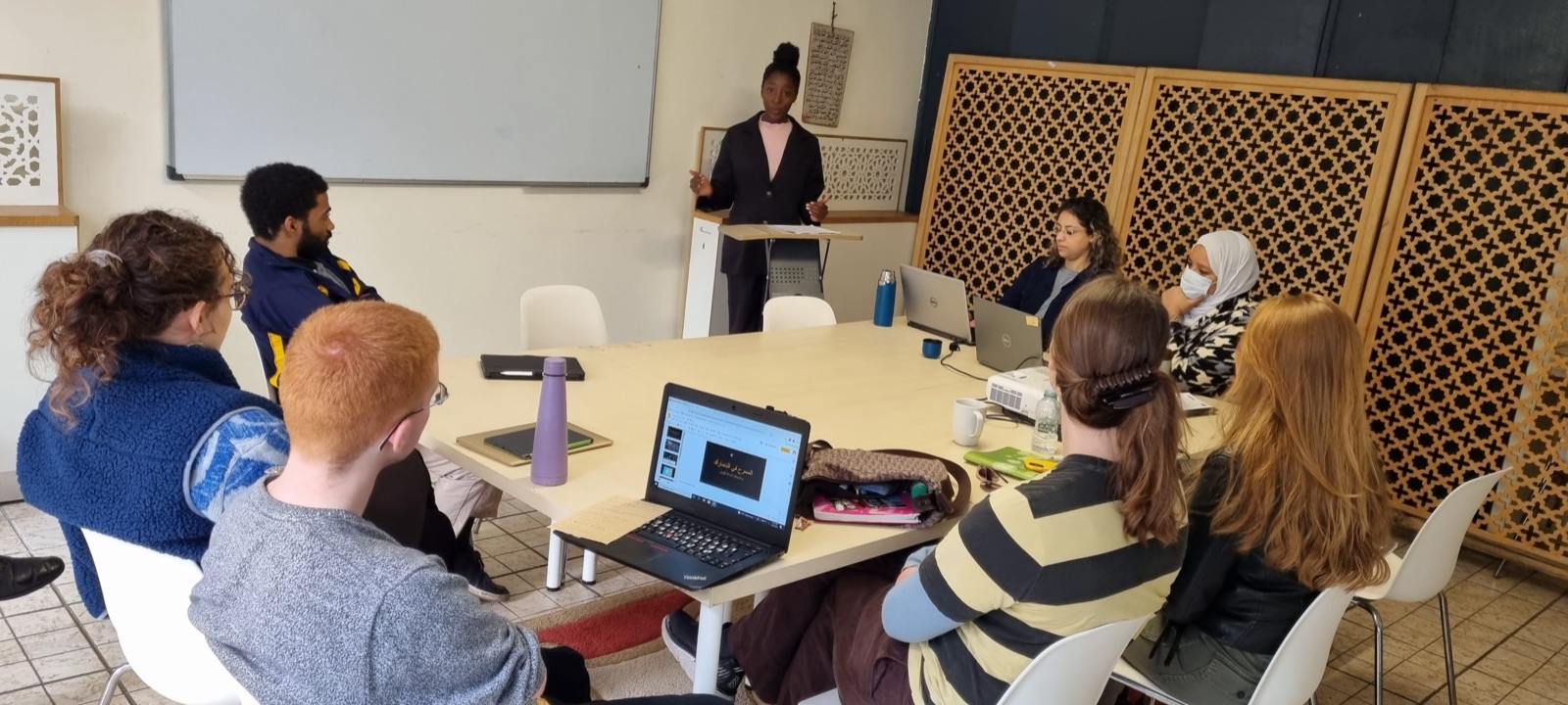 A student presents to her group around a long table, with laptops open and carved wooden screens behind them