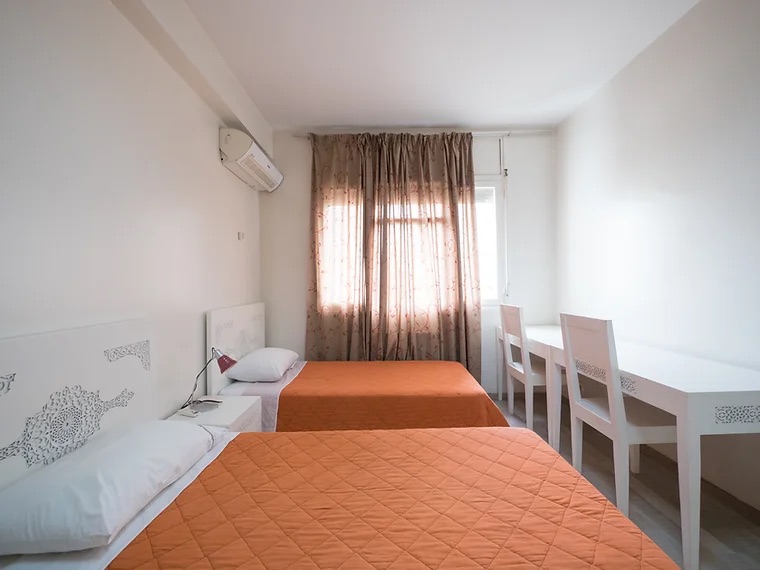 A bright twin room in the student residence, with orange bedspreads, carved white headboards and two desks by the window