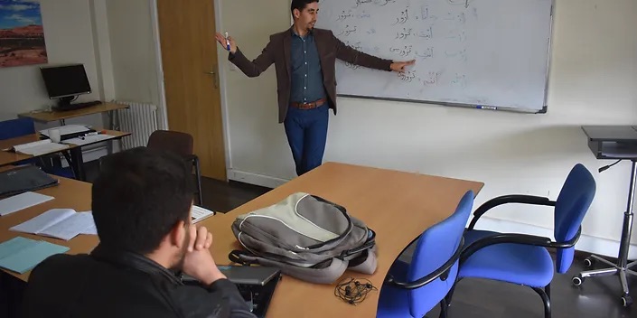 A teacher points to Arabic words on the whiteboard while a student listens from the table